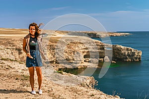 Attractive young woman stands on a high cliff overlooking the rocky cliffs arches on the beach and turquoise sea water on the coas