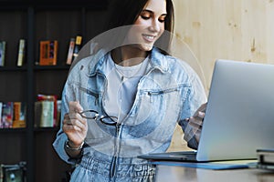 Attractive young woman stands near the window, holds smartphone in her hand, glasses for vision and works on laptop.