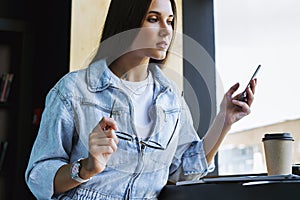 Attractive young woman stands near the window, holds smartphone in her hand, glasses for vision and works on laptop.