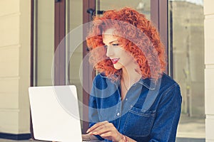 Attractive young woman student in profile, working on laptop, browsing, sitting outdoors. Thoughtful daydreaming girl with red