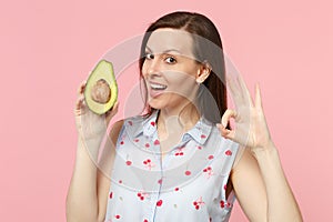 Attractive young woman in summer clothes showing OK gesture, hold fresh ripe green avocado fruit isolated on pink pastel