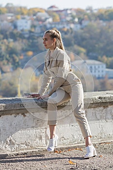 Attractive young woman in sunglasses and overalls sits on retaining wall in public park