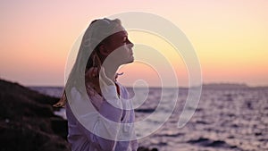 attractive young woman at sunset on background of ocean or sea in white shirt and black top looks into distance, then