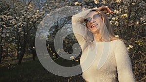 Attractive young woman in sweater, jeans is dance and spinning among blossom apple tree. Sunset, spring orchard and