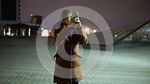 Attractive young woman taking pictures on a smartphone during walk on streets at night town. Slow motion
