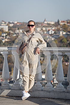 Attractive young woman in tinted sunglasses and overalls posing leaning on balustrade crossed legs and looking at camera