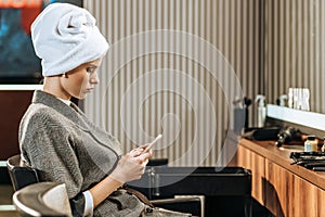 Attractive young woman with towel on head sitting and using smartphone