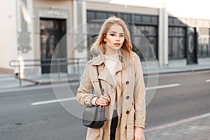 Attractive young woman in trendy spring coat in white t-shirt with stylish black leather handbag posing in the city outside.