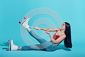 Attractive young woman tying roller skate and smiling while sitting against blue background