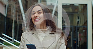 Attractive young woman using phone walking near big modern office building. Chatting with friends, girl using cellphone