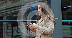 Attractive young woman using phone walking near big modern office building. Chatting with friends, girl using cellphone