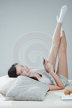 Attractive young woman using smartphone while lying on bed and smiling