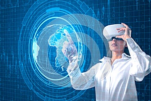 Attractive young woman with VR glasses using holographic screen with digital globe interface and business chart on blurry blue