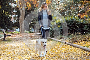 Attractive young woman walking with her lovely golden retriever dog while using her smart phone in the park in autumn