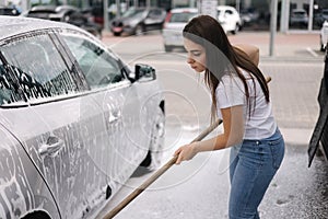 Attractive young woman washing her car with shampoo and brushes. Female washes automobile with foam and water outside on