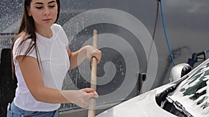 Attractive young woman washing her car with shampoo and brushes. Female washes automobile with foam and water outside on