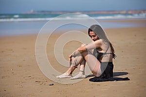 Attractive young woman, wearing black swimsuit and sarong, lonely, quiet and sad, sitting on the shore of the beach. Concept