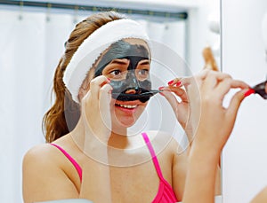 Attractive young woman wearing pink top and white headband, removing black mask treatment from face using hands, looking