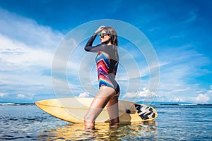 attractive young woman in wetsuit and sunglasses with surfboard posing in ocean at Nusa dua Beach