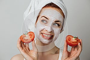 Attractive young woman with white cosmetic mask on her face holding sliced fresh tomato.
