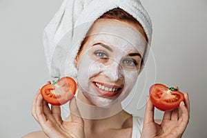 Attractive young woman with white cosmetic mask on her face holding sliced fresh tomato.