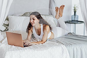 An attractive young woman in a white fitness suit is lying on the bed and typing on a laptop in the bedroom. A charming curly-