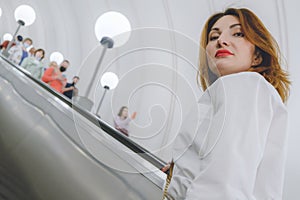 Attractive young woman in a white shirt and with bright red lips rides up the escalator in the subway