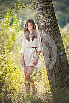 Attractive young woman in white short dress posing near a tree in a sunny summer day. Beautiful girl enjoying the nature