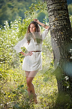 Attractive young woman in white short dress posing near a tree in a sunny summer day. Beautiful girl enjoying the nature