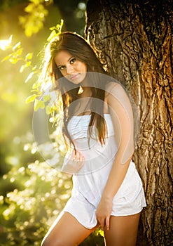 Attractive young woman in white short dress posing near a tree in a sunny summer day. Beautiful girl enjoying the nature