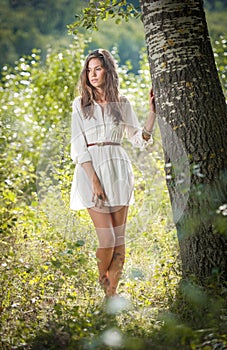 Attractive young woman in white short dress posing near a tree in a sunny summer day. Beautiful girl enjoying the nature