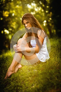 Attractive young woman in white short dress sitting on grass in a sunny summer day. Beautiful girl enjoying the nature