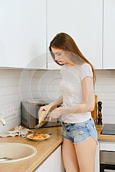 Attractive young woman in white t-shirt and denim shorts grates parmesan on a hand grater over a plate of pasta