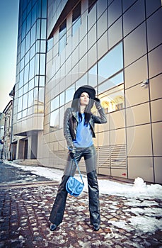 Attractive young woman in a winter fashion shot. Beautiful fashionable young girl in black leather with big hat and blue handbag