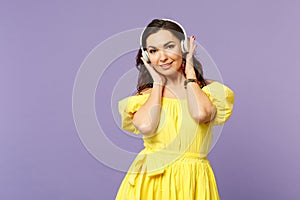 Attractive young woman in yellow dress looking camera, listen music, putting hands on headphones isolated on pastel