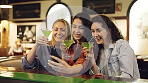 Attractive young women are taking selfie with cocktails in bar. Cheerful girls are posing, laughing and clanging glasses