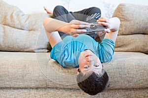 Attractive youngster guy wearing blue t-shirt lying on his back on couch and holding smartphone in hands, browsing social media