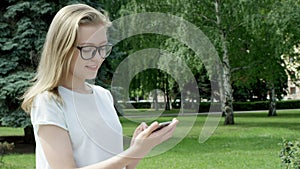 Attractive youthful female chatting absorbedly using smartphone during a sunny day. Blonde girl stands in park in front