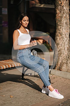 Attrective brunette sitting alone on the bench in centre of the city. Beautiful young woman drinking coffe outdoors. End