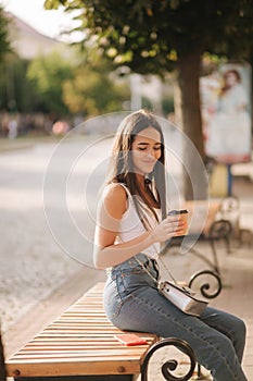 Attrective brunette sitting alone on the bench in centre of the city. Beautiful young woman drinking coffe outdoors. End