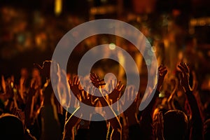 Audience with hands raised at a music festival and lights streaming down from above the stage.