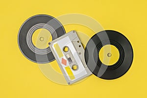 Audio cassette with magnetic tape and vinyl discs on a yellow background.