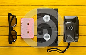 Audio cassette, vhs, 3d glasses, hipster film camera on a yellow wooden background. Retro devices from 80s. Top view