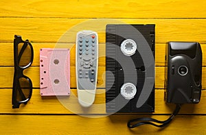 Audio cassette, vhs, 3d glasses, tv remote, hipster film camera on a yellow wooden background. Retro devices from 80s. Top view