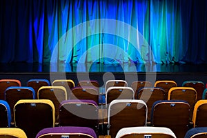 The auditorium in the theater. Blue-green curtain on the stage. Multicolored spectator chairs. Lighting equipment