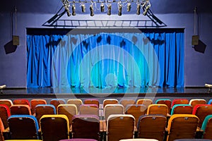 The auditorium in the theater. Blue-green curtain on the stage. Multicolored spectator chairs. Lighting equipment