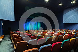 The auditorium in the theater. Blue-green curtain on the stage. Multicolored spectator chairs. Lighting equipment