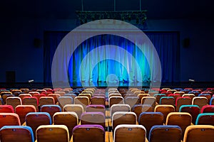 The auditorium in the theater. Blue-green curtain on the stage. Multicolored spectator chairs. Lighting equipment