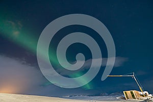 Aurora borealis with full moon lit clouds, glow of houses, construction crane and totally snowy ground with bluish starry sky in