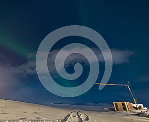 Aurora borealis with full moon lit clouds, glow of houses, construction crane and totally snowy ground with bluish starry sky in